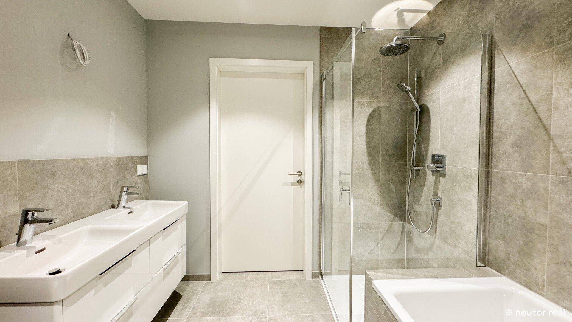 Helles Badezimmer mit Doppelwaschbecken, moderner Dusche und Zugang zu weiteren Räumen.