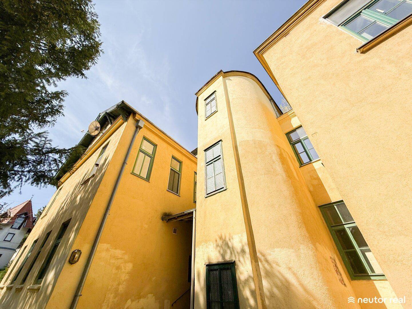 Detailansicht der gelben Fassade mit runden Erkern und grünen Fenstern unter blauem Himmel.