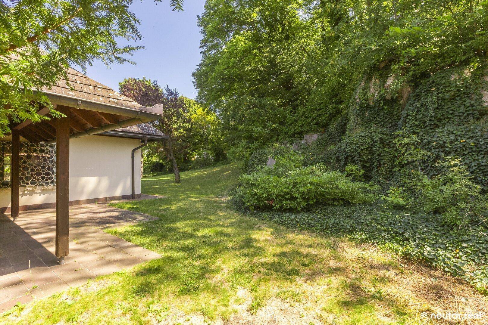 Sonniger Garten mit Rasen, Bäumen und einer überdachten Terrasse am Haus.