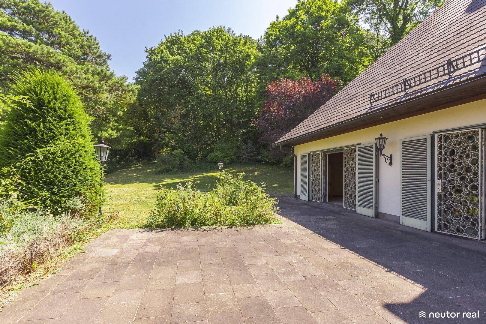 Geräumige Terrasse mit Steinplatten und Laternen, umgeben von üppigem Grün und Bäumen.