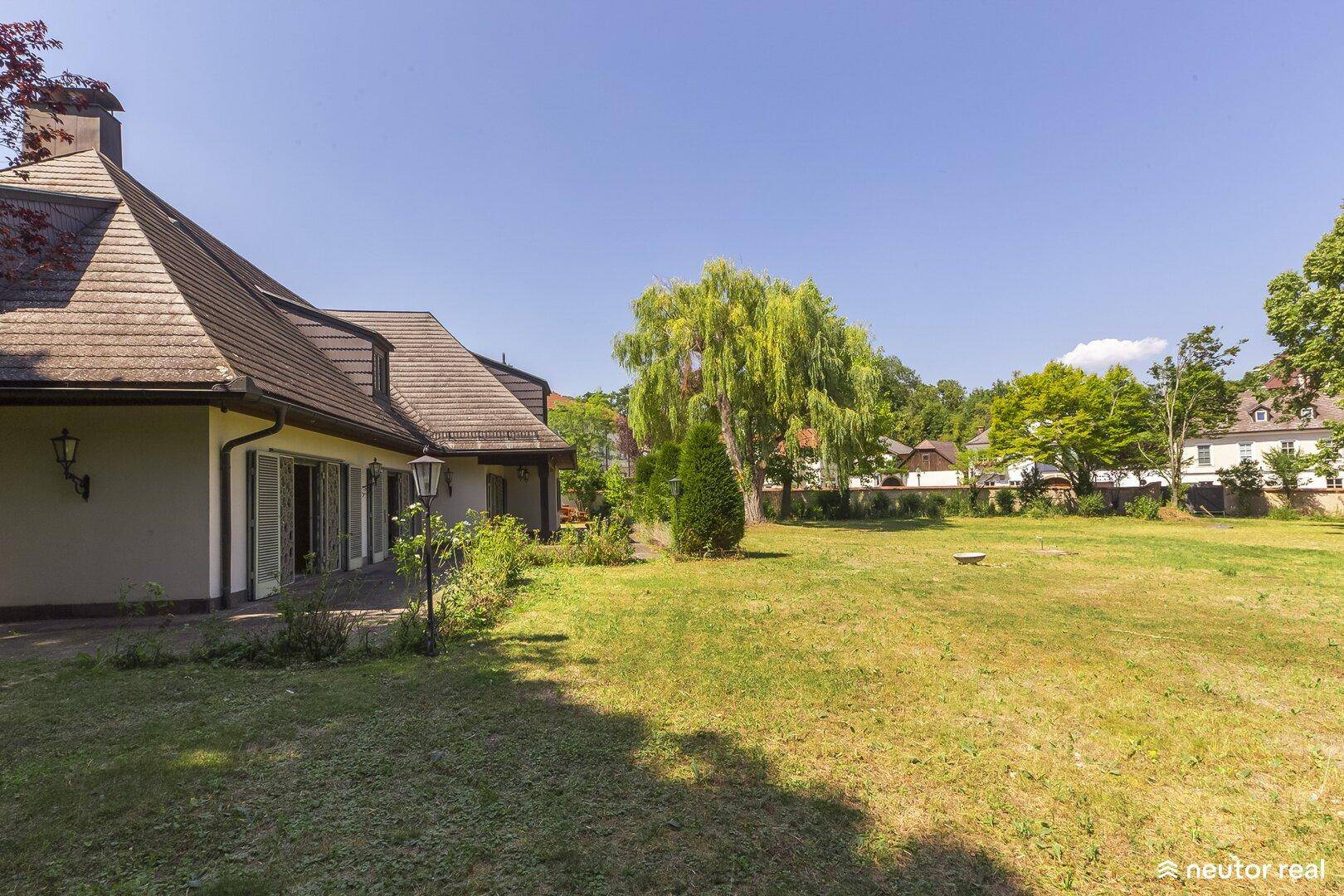 Seitenansicht des Hauses mit weitläufigem Rasen und altem Baumbestand unter blauem Himmel.