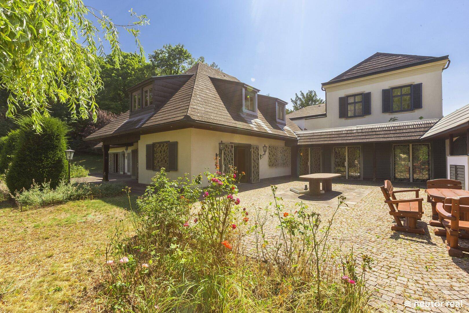 Charmante Terrasse mit Kopfsteinpflaster, blühenden Pflanzen und Blick auf das Landhaus.