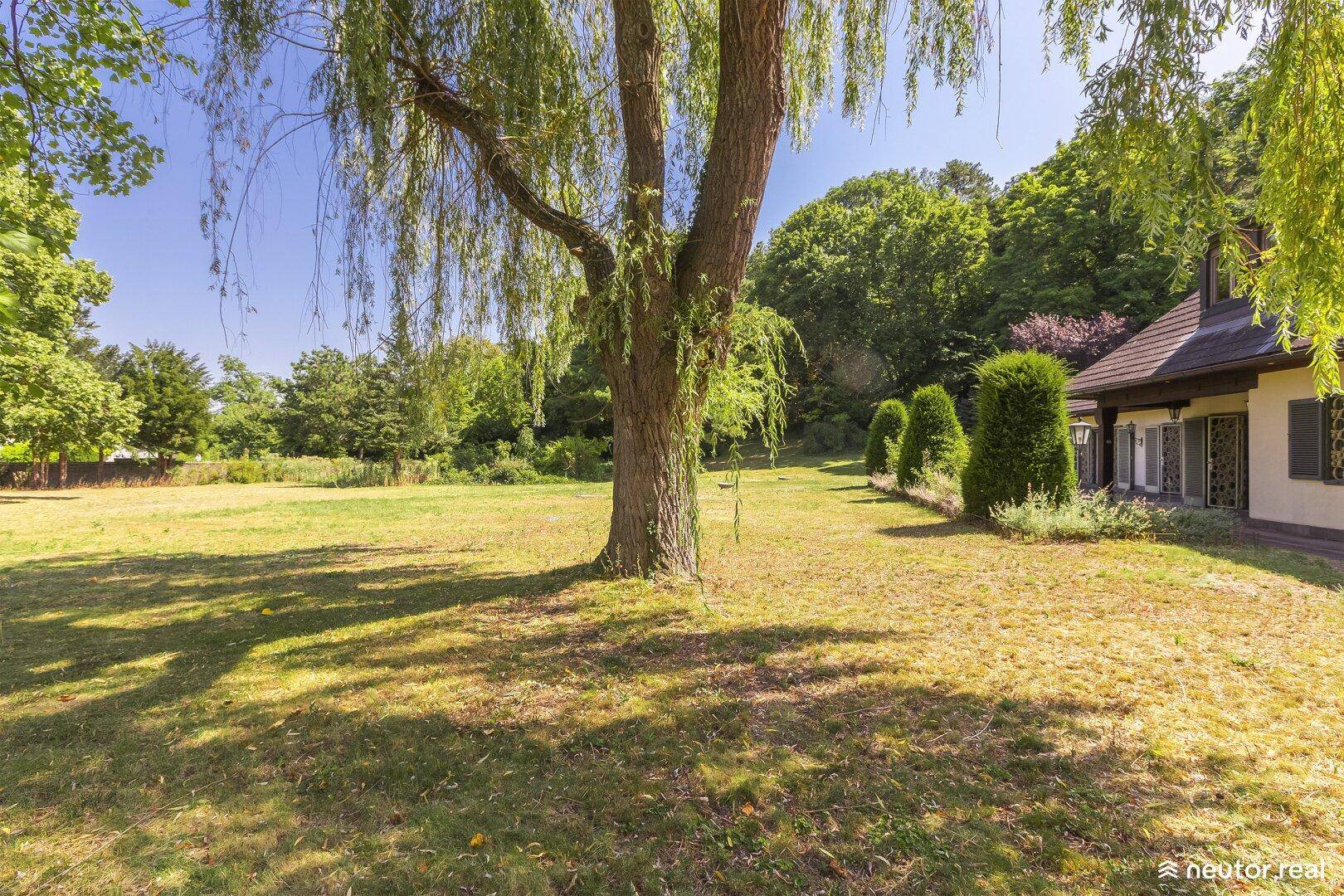 Weitläufiger Garten mit altem Baumbestand und Blick auf das charmante Landhaus unter blauem Himmel.