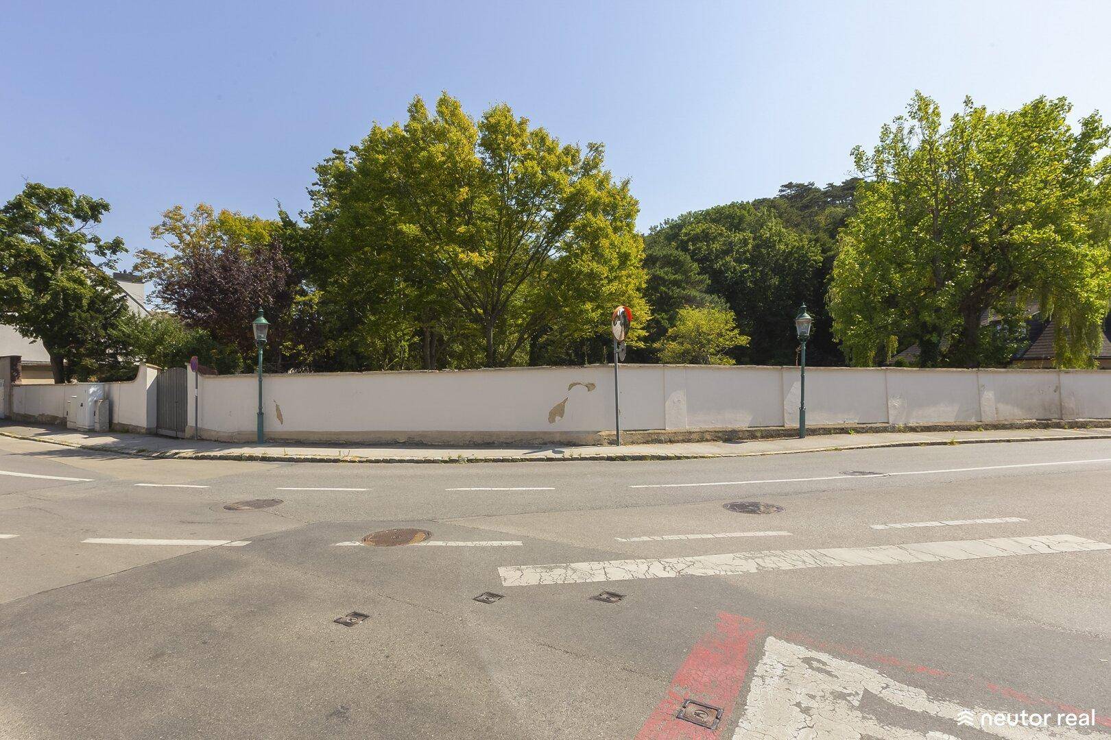 Straßenansicht mit langer weißer Mauer, die das Grundstück umgibt, und üppigem Baumbestand.