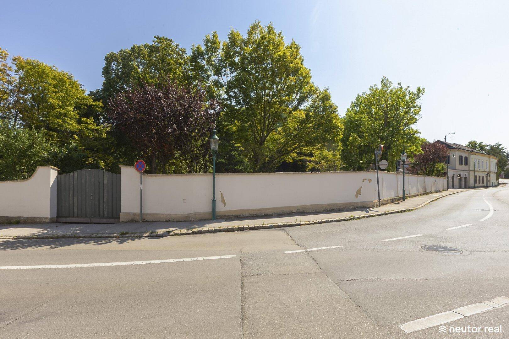 Straßenansicht mit langer weißer Mauer, Holztor und üppigem Baumbestand im Hintergrund.