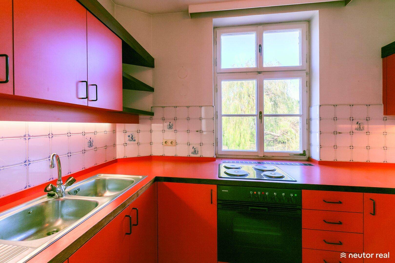 Rote Küche mit weißen Fliesen, Spüle, Herd und Blick ins Grüne durch das Fenster.