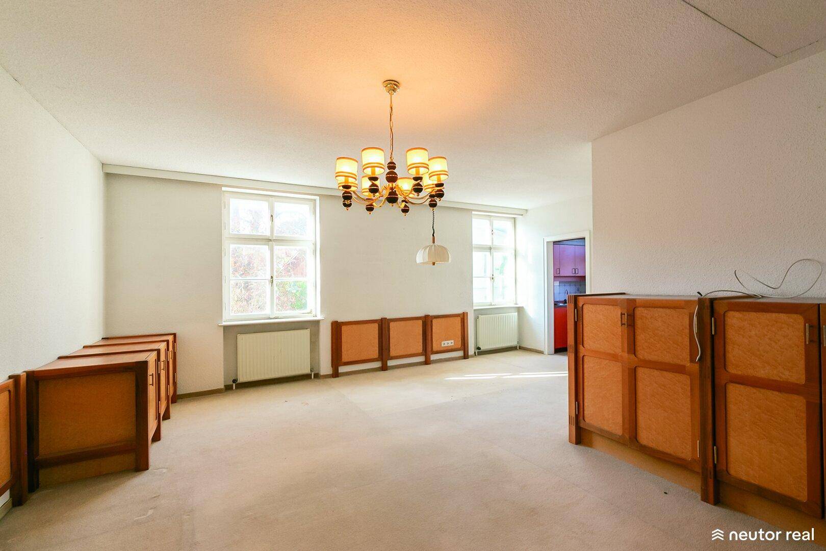 Geräumiges, leeres Wohnzimmer mit zwei Fenstern, Kronleuchter und Holzmöbeln.