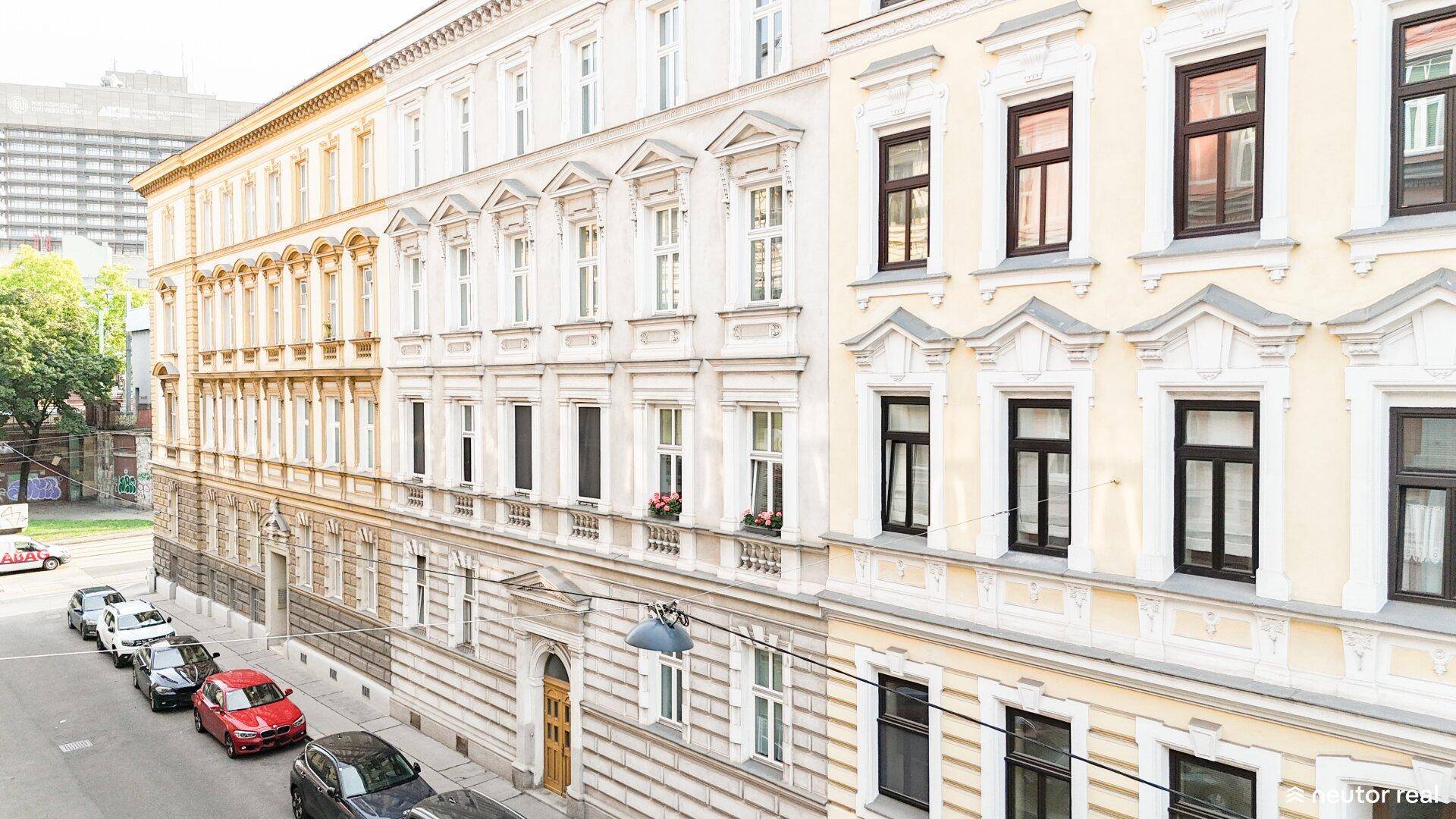 Historische Gebäudefassaden entlang einer Straße mit parkenden Autos und Bäumen im Hintergrund.