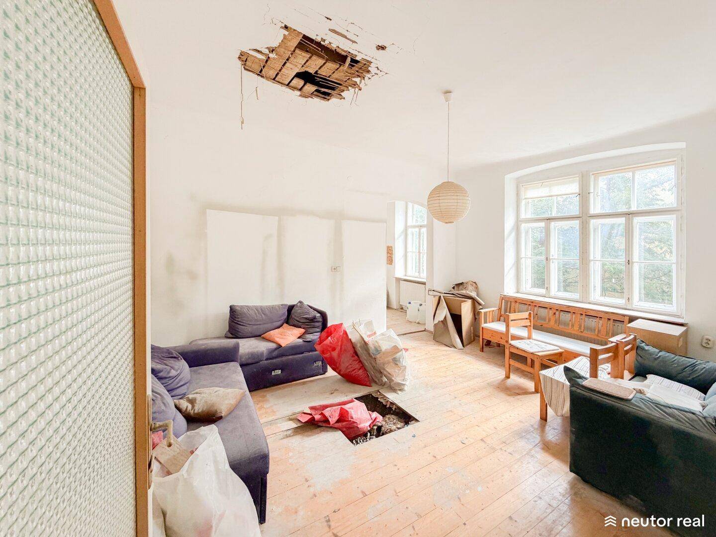 Geräumiges Wohnzimmer mit Holzboden, großen Fenstern und sichtbaren Renovierungsarbeiten an der Decke.