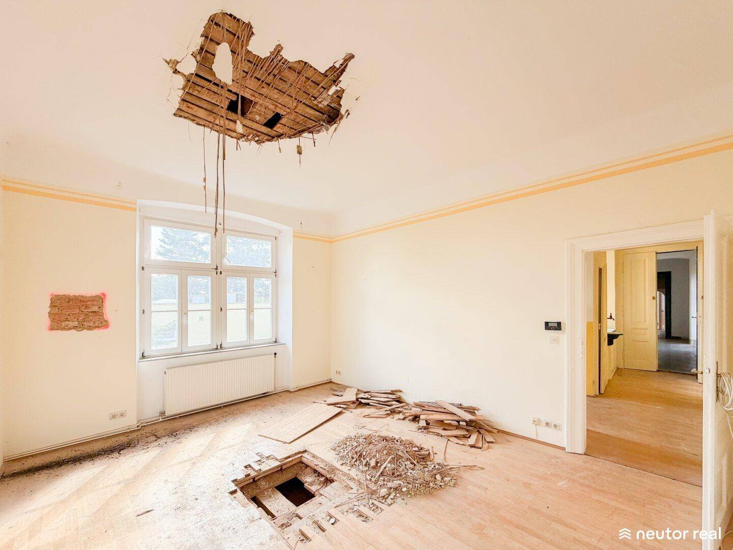 Geräumiges Wohnzimmer mit Holzboden, großen Fenstern und sichtbaren Schäden an Decke und Boden.