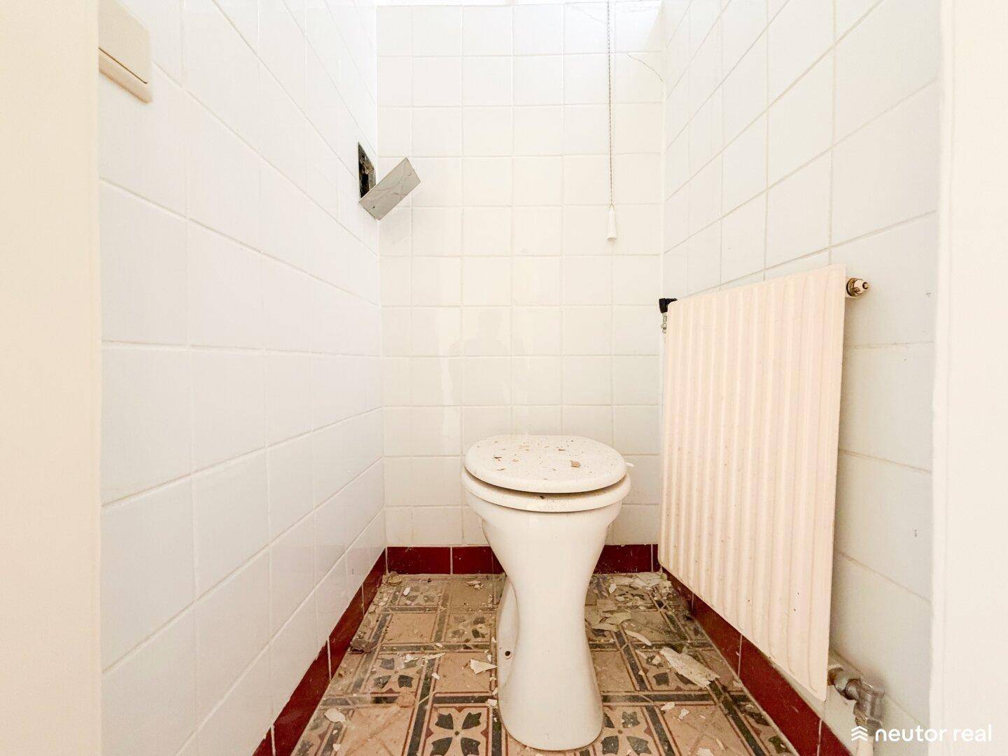 Kleines WC mit weiß gefliesten Wänden, Stand-WC und Heizkörper, in renovierungsbedürftigem Zustand.