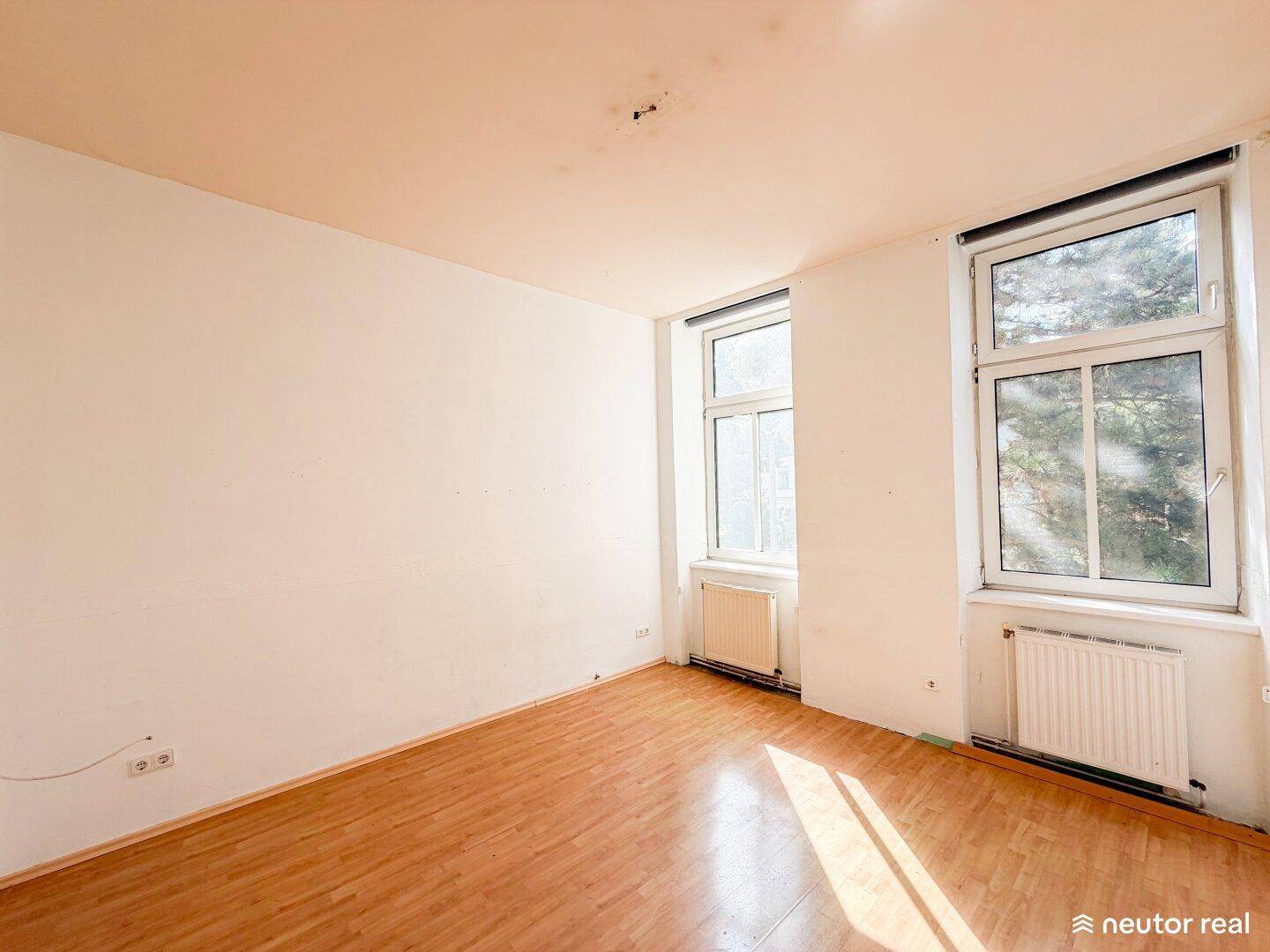 Helles Wohnzimmer mit Laminatboden und zwei großen Fenstern, die viel Tageslicht hereinlassen.