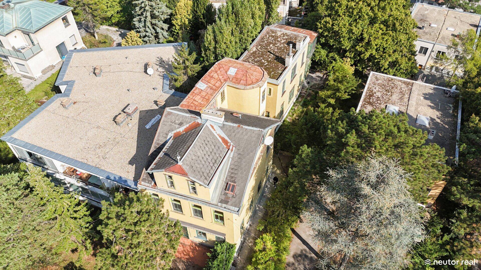 Vogelperspektive einer gelben Villa mit verschiedenen Dachformen, eingebettet in grüne Umgebung.