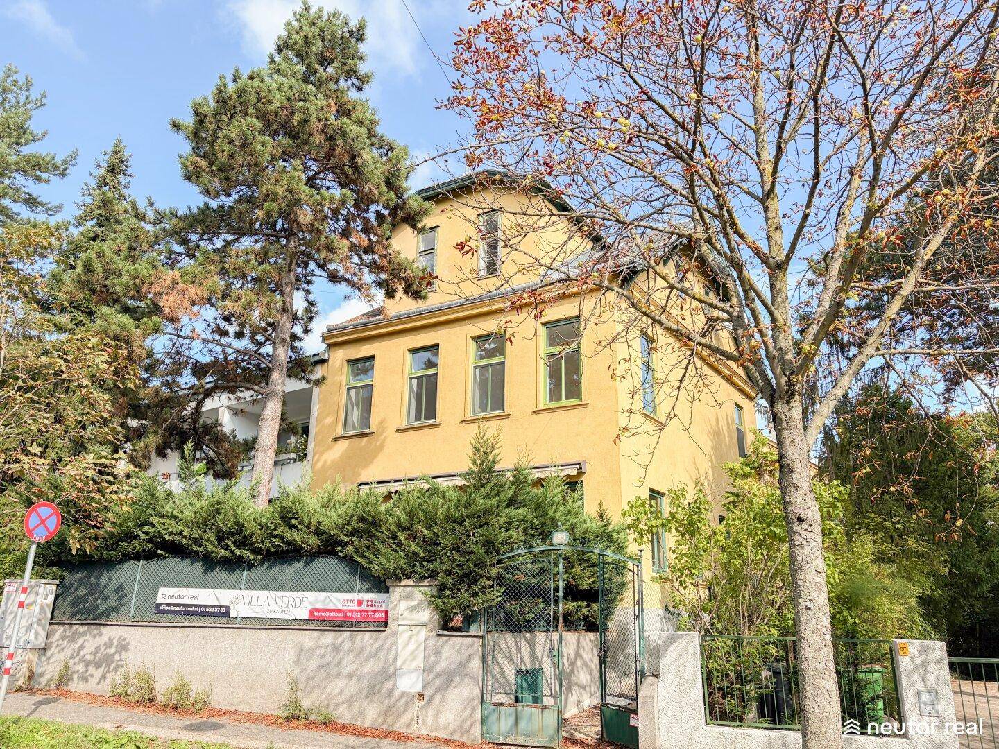 Gelbe Fassade eines Altbaus mit grünen Fensterrahmen und umgebender Vegetation.
