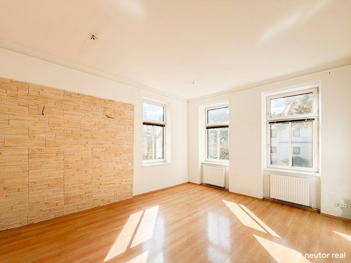 Geräumiges Wohnzimmer mit Holzboden, drei Fenstern und einer dekorativen Wandverkleidung.