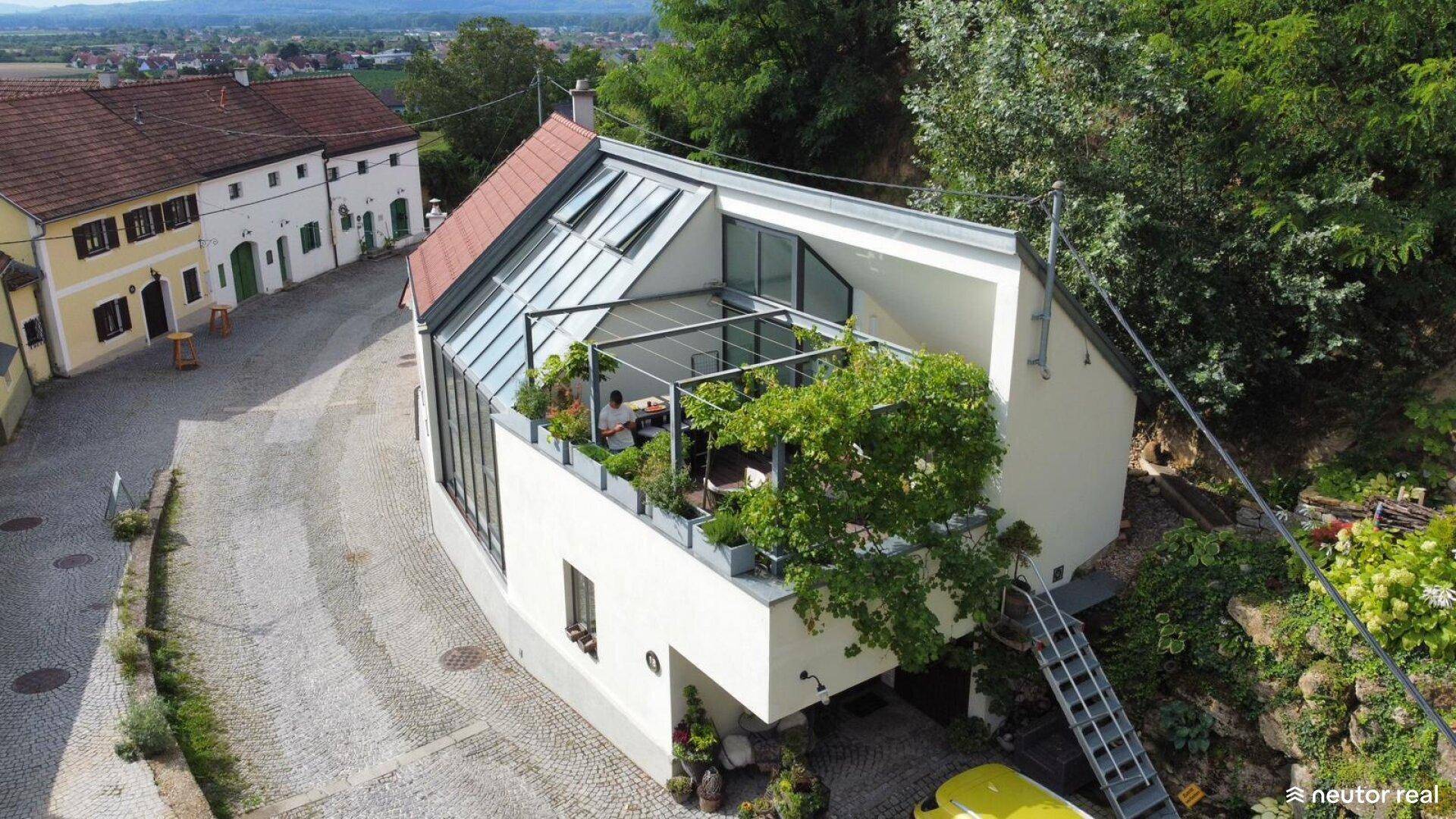 Moderne Architektur mit großer Dachterrasse und Glasdach, umgeben von traditionellen Gebäuden und viel Grün.