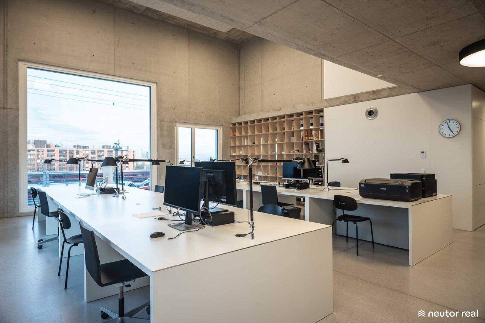 Offener Bürobereich mit langen Schreibtischen, Computern und großen Fenstern mit Blick auf die Stadt.