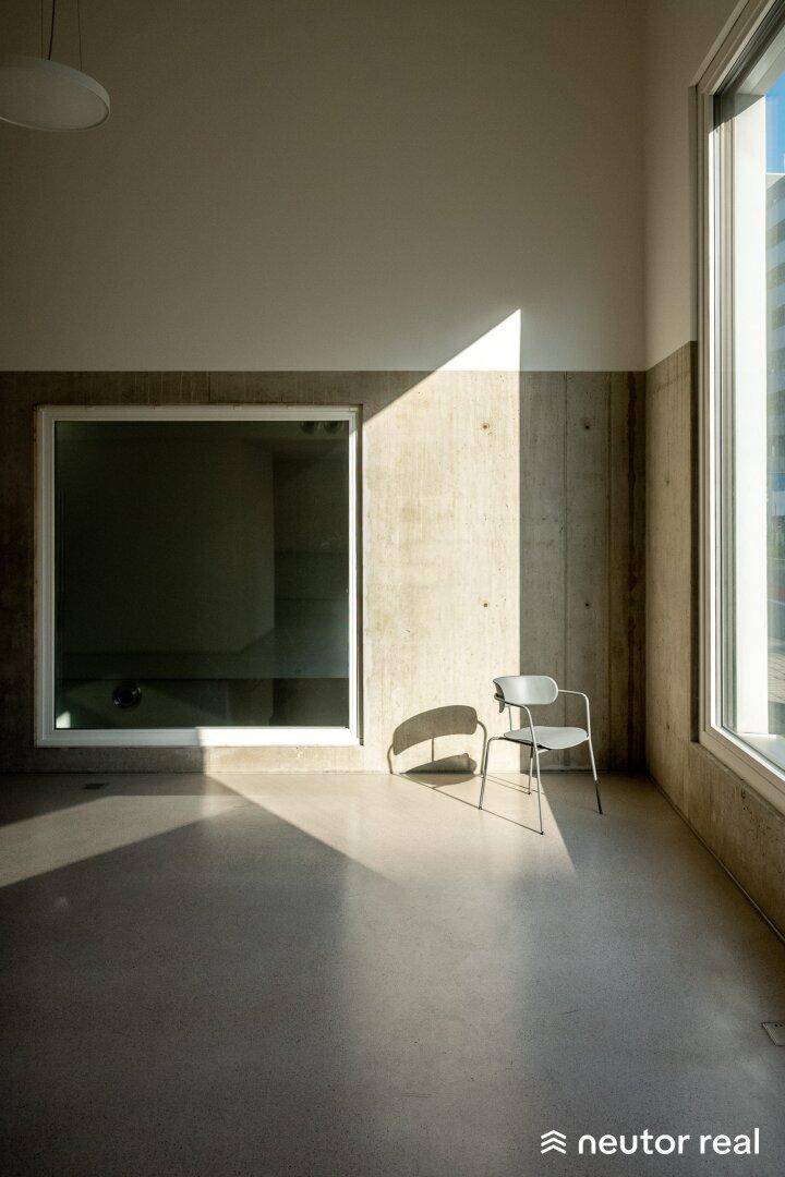 Heller Raum mit Sichtbetonwänden, großen Fenstern und starkem Lichteinfall, der Schatten wirft.