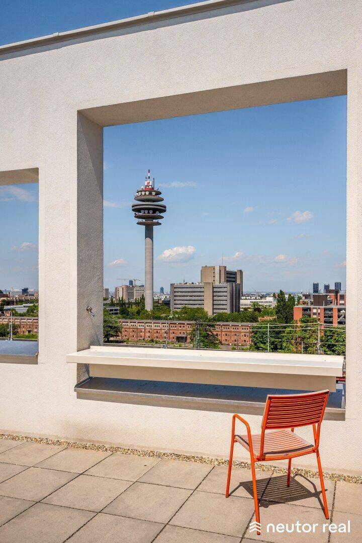 Sonniger Terrassenbereich mit orangefarbenem Stuhl und weitem Blick über die Stadt mit Fernsehturm.