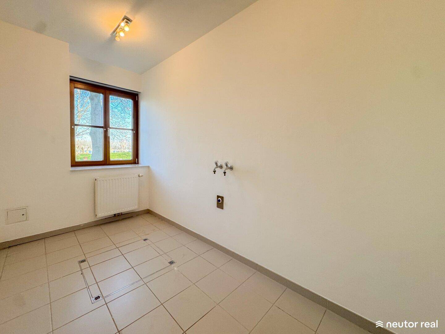 Leerer Raum mit Fliesenboden, Fenster und Anschlüssen für Wasser, ideal als Hauswirtschaftsraum.