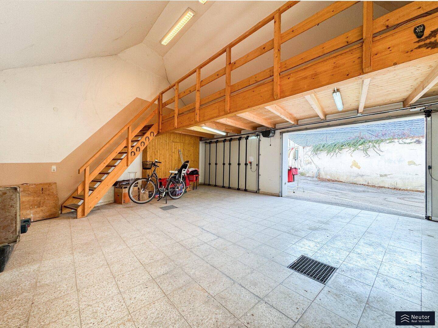Große Garage mit Fliesenboden, Holztreppe und einem zusätzlichen Lagerbereich im Obergeschoss.