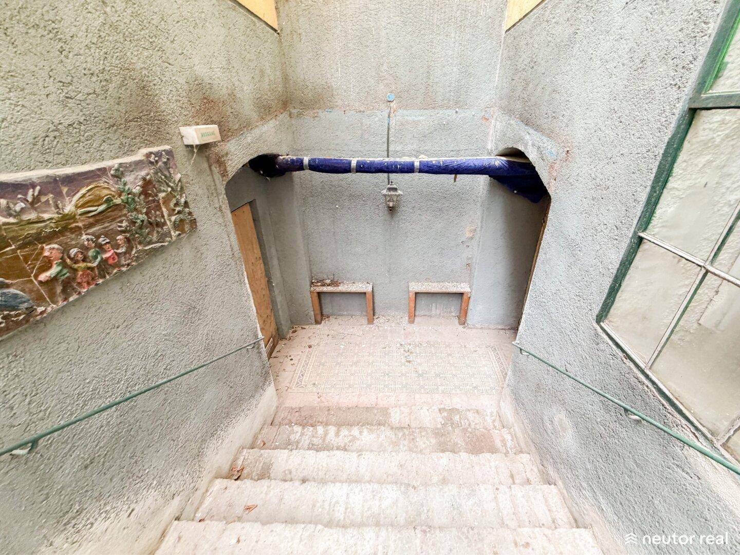 Treppenabgang in den Keller mit rauen Wänden, Fliesenboden und einem blauen Rohr.