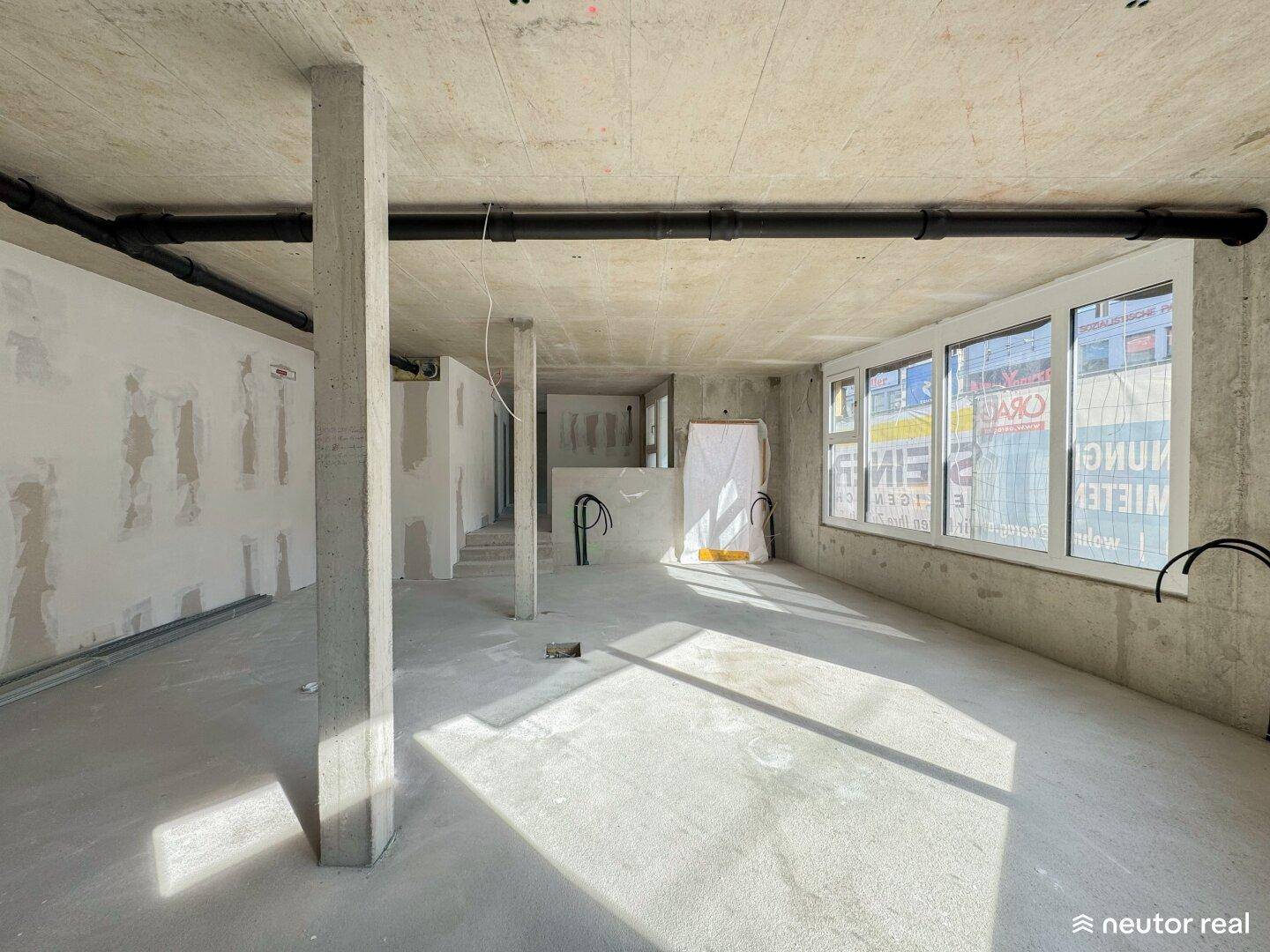 Ein weitläufiger, unfertiger Innenraum mit Betonwänden und -decken, großen Fenstern und sichtbaren Rohren im Rohbauzustand.