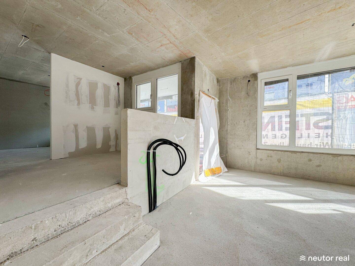 Ein unfertiger Raum mit Betonwänden, großen Fenstern und einer Treppe im Rohbauzustand, bereit für den Ausbau.