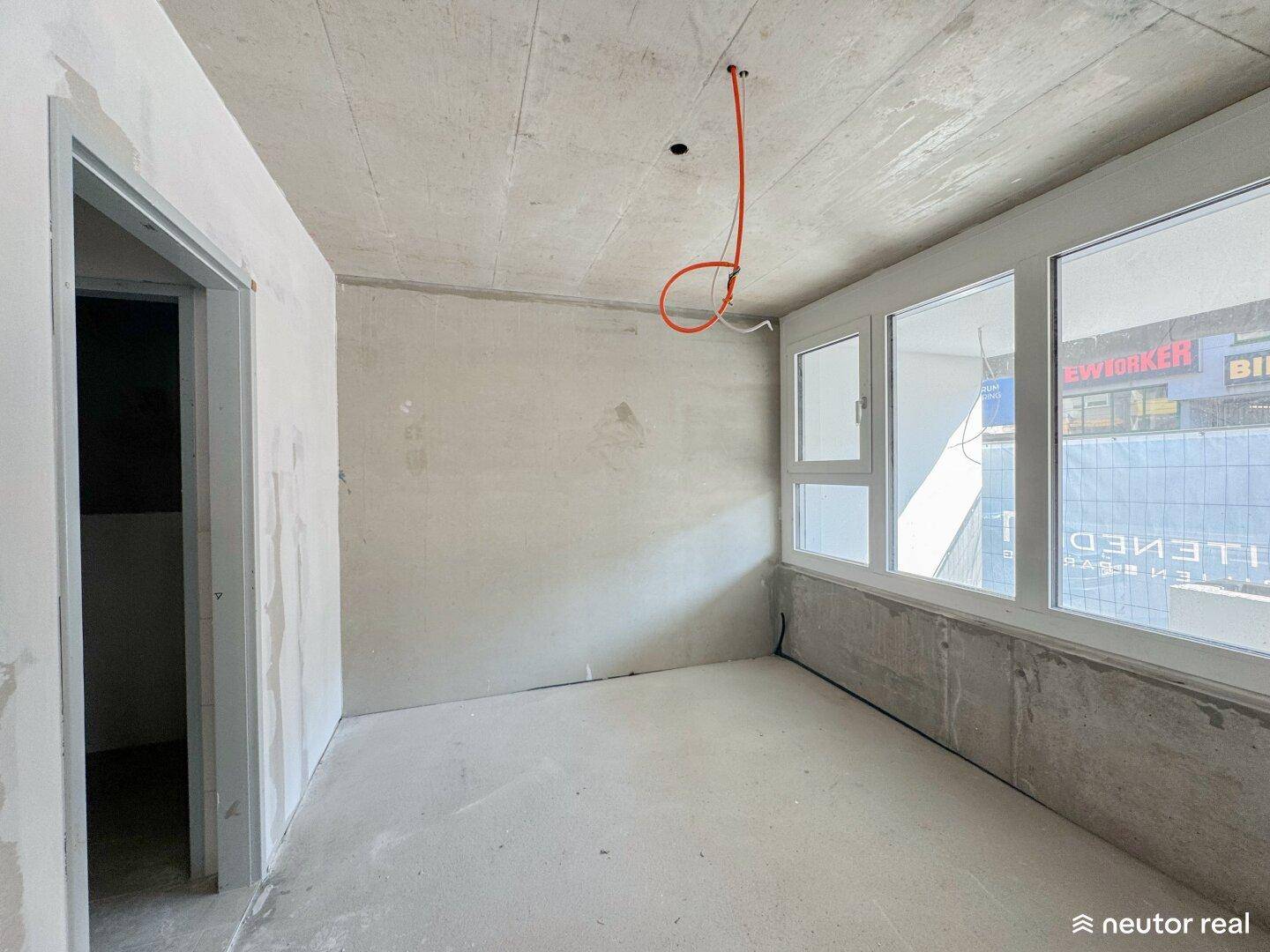 Ein kleinerer, unfertiger Raum mit Betonwänden, einem Fenster und sichtbaren Elektrokabeln an der Decke im Rohbauzustand.
