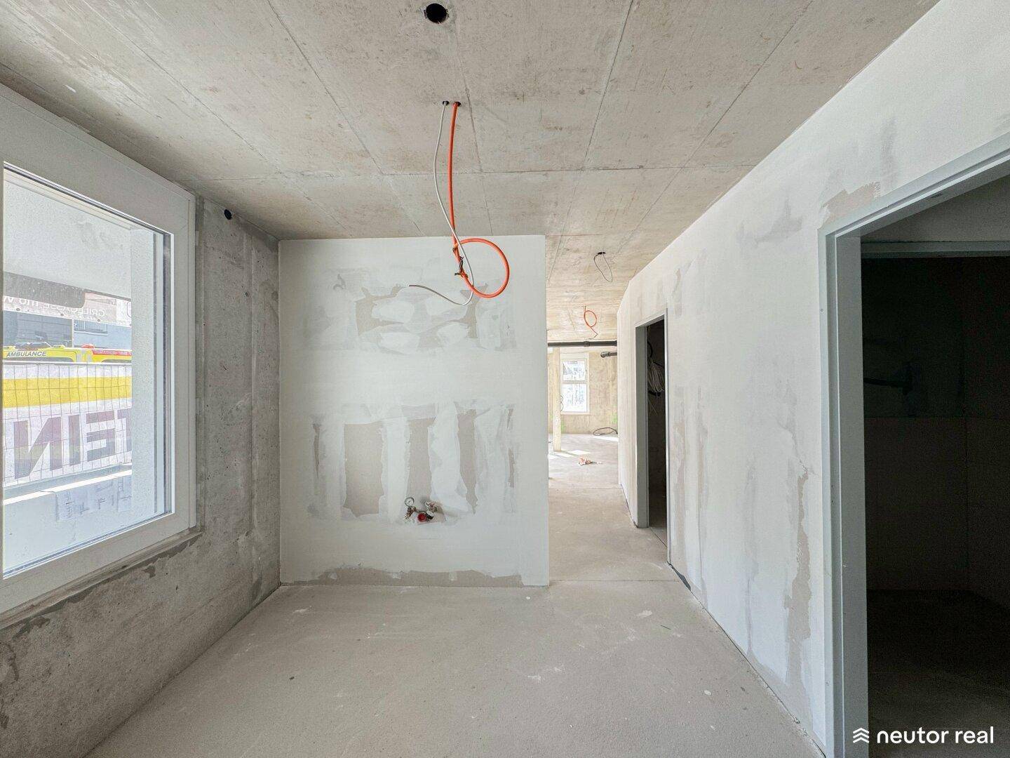 Ein unfertiger Innenbereich mit Betonwänden, einem Fenster und sichtbaren Elektrokabeln an der Decke im Rohbauzustand.