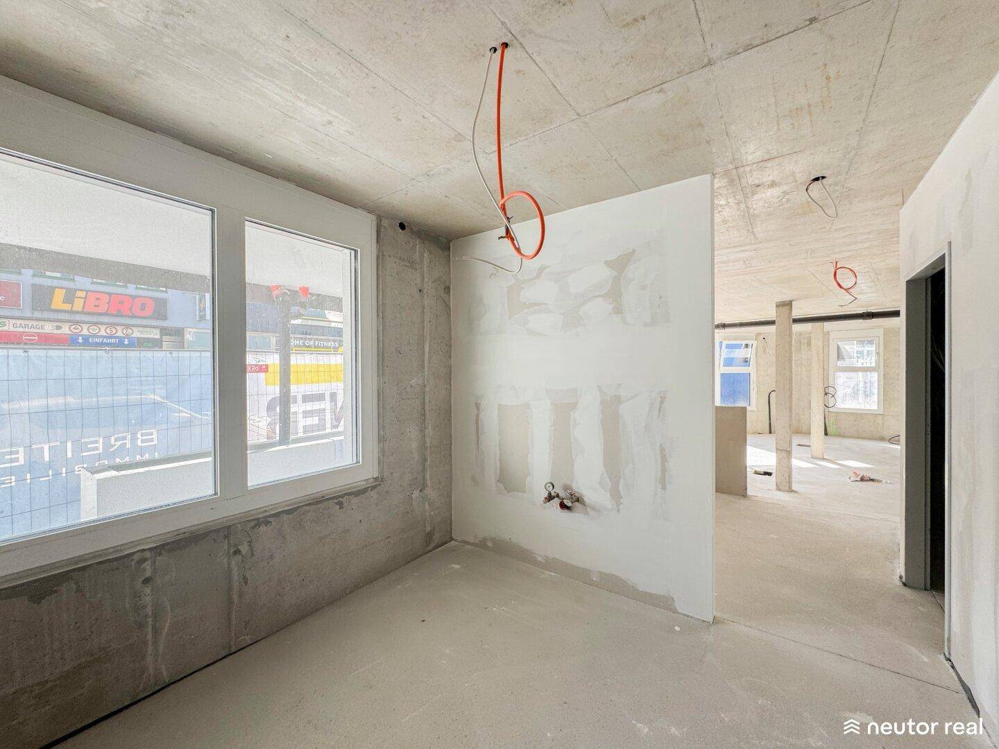 Ein unfertiger Raum mit Betonwänden, einem Fenster und sichtbaren Elektrokabeln an der Decke im Rohbauzustand.