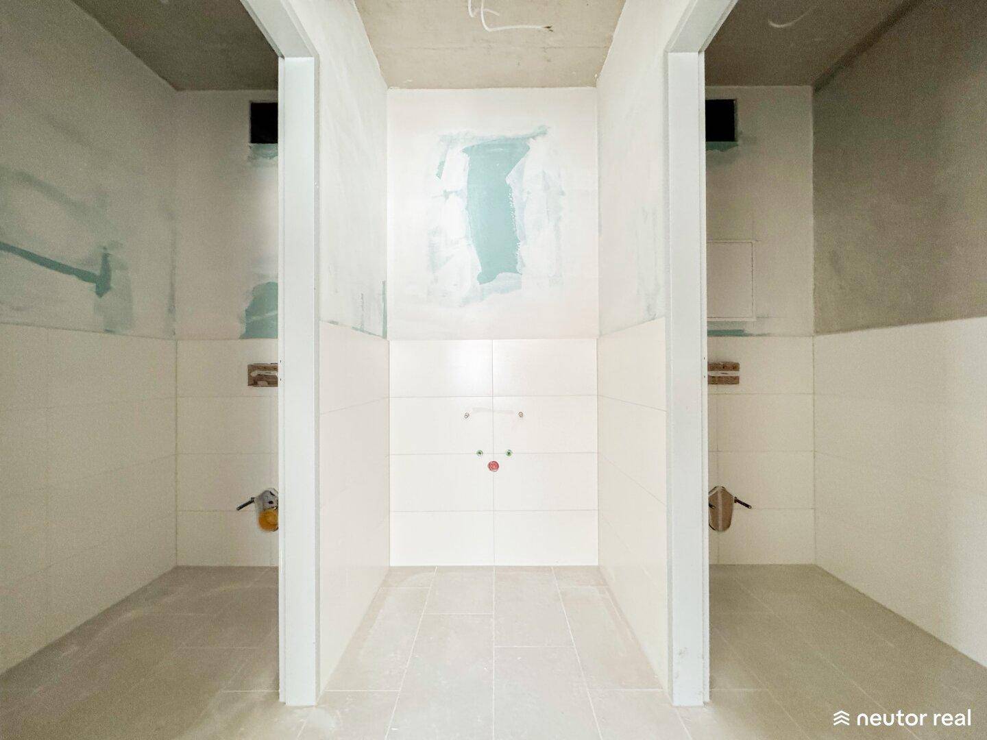 Ein im Rohbau befindlicher Sanitärbereich mit gefliesten Wänden und vorbereiteten Anschlüssen für die Installation.