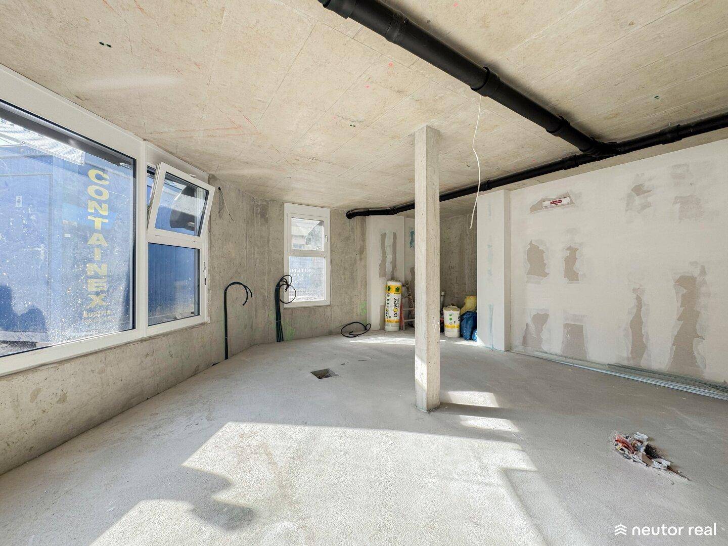 Ein unfertiger Raum mit Betonwänden, großen Fenstern und sichtbaren Rohren an der Decke im Rohbauzustand.