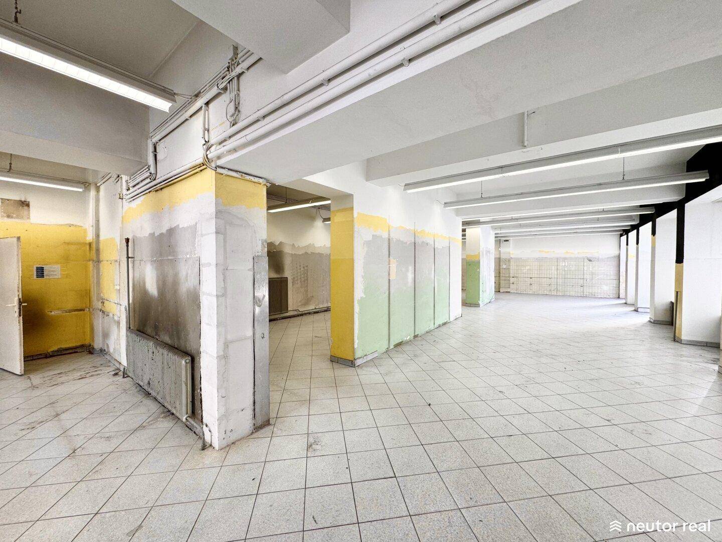 Weiterer Blick in den weitläufigen Gewerberaum mit Fliesenboden, sichtbaren Leitungen und teilweise gestrichenen Wänden.
