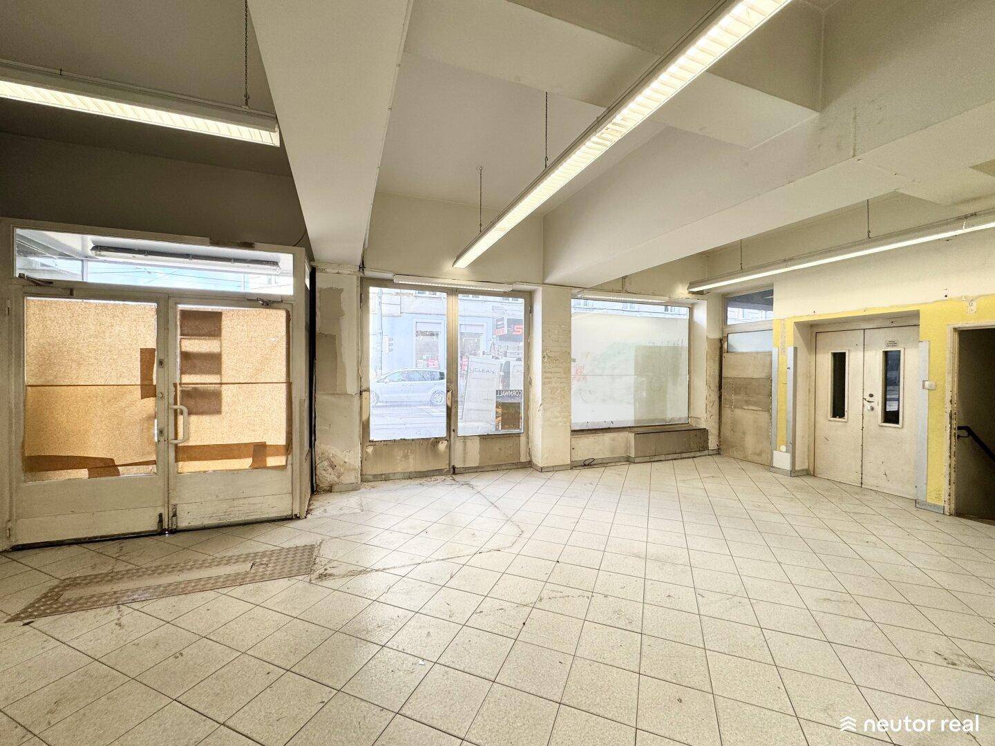 Eingangsbereich des Gewerberaums mit großen Glastüren und Schaufenstern zur Straße hin, auf Fliesenboden.