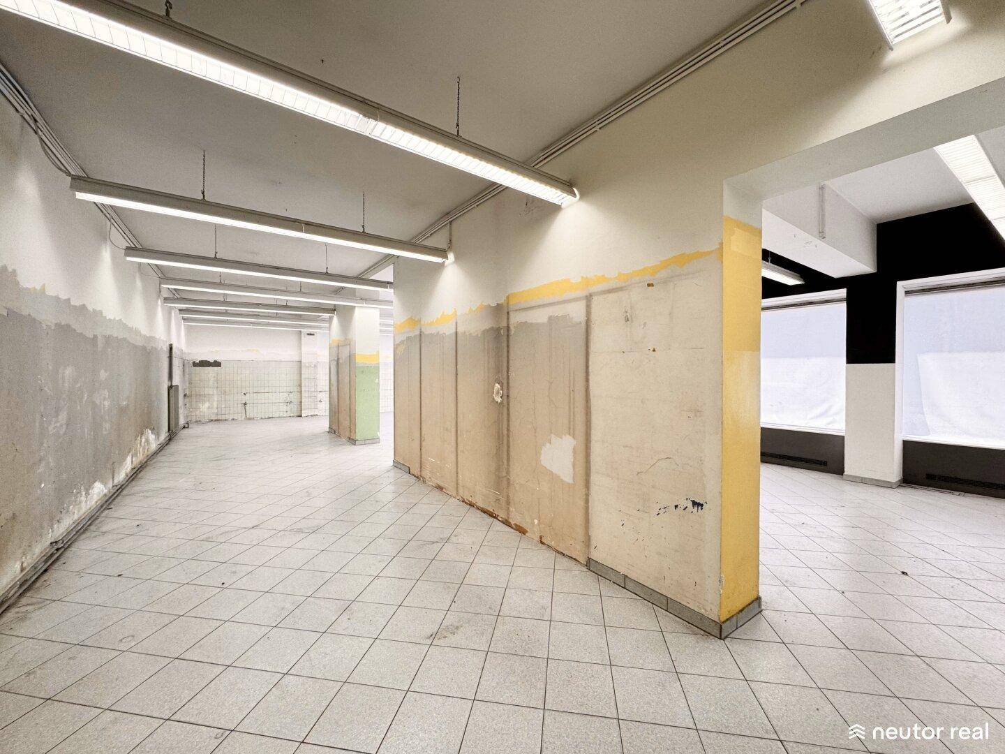 Ansicht des Gewerberaums mit Fliesenboden, langen Leuchtstoffröhren und einer Wand mit sichtbaren Putzspuren und gelber Farbe.