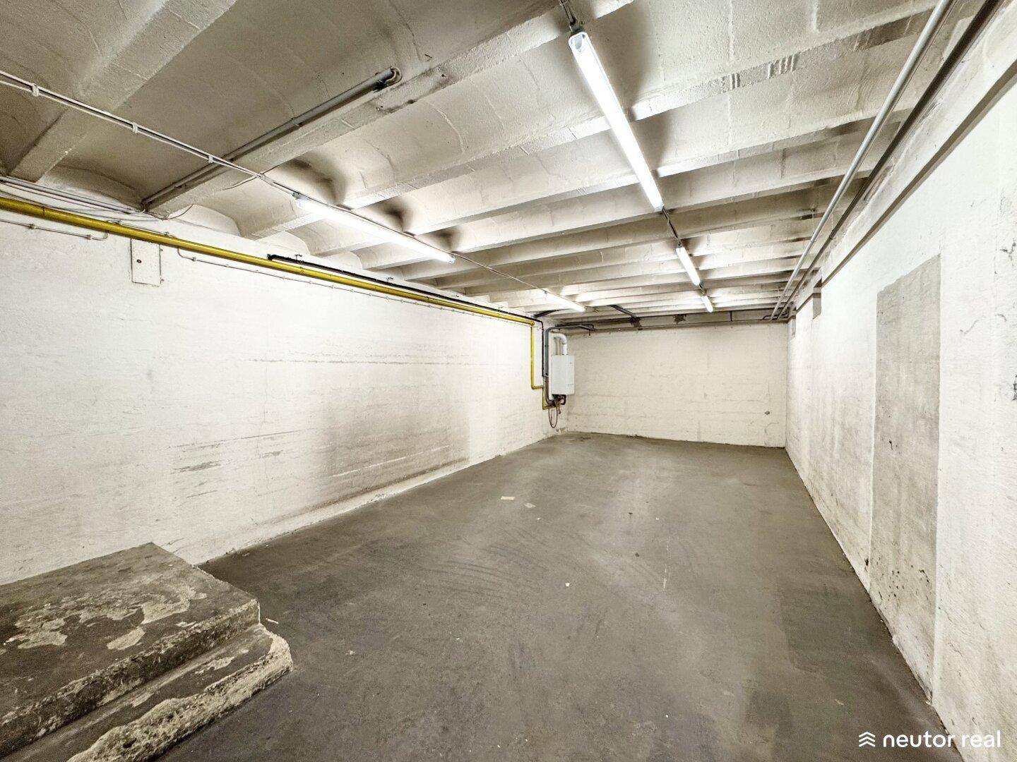 Kellerraum mit Betonboden, weiß gestrichenen Ziegelwänden und sichtbaren Leitungen an der Decke.