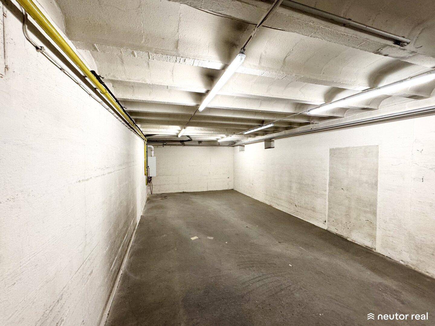 Leerer Kellerraum mit Betonwänden, Deckenleuchten und sichtbaren Leitungen, der viel Stauraum bietet.