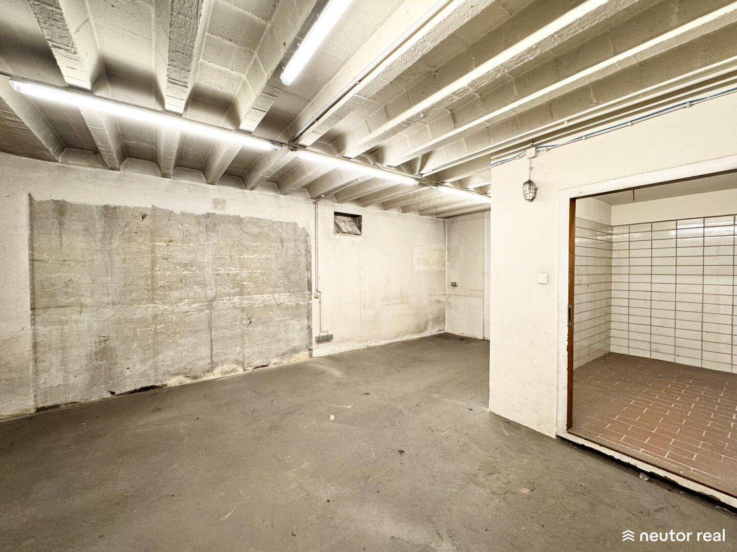 Kellerraum mit Betonboden und einer offenen Tür zu einem gefliesten Bereich, die Wände zeigen Gebrauchsspuren.