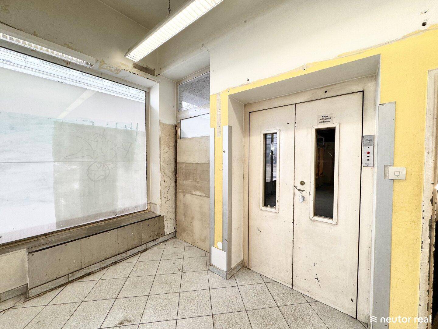 Alter Flur mit Fliesenboden, einem Aufzug und einem großen Fenster, das Tageslicht hereinlässt.