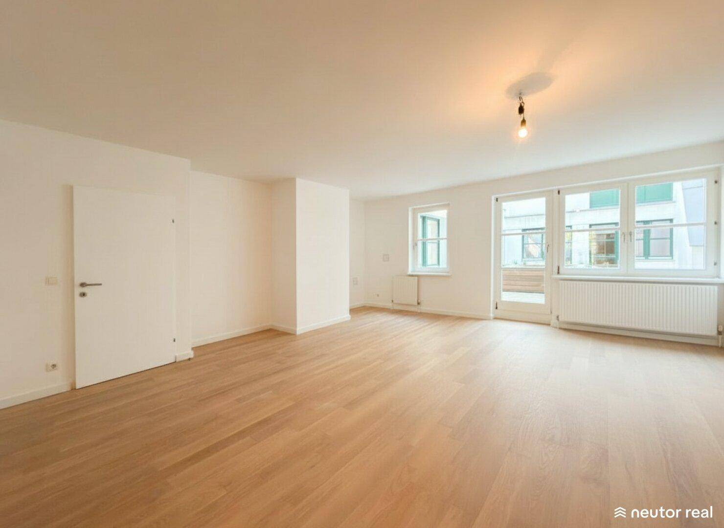Heller, leerer Raum mit Holzboden, großen Fenstern und Zugang nach draußen.