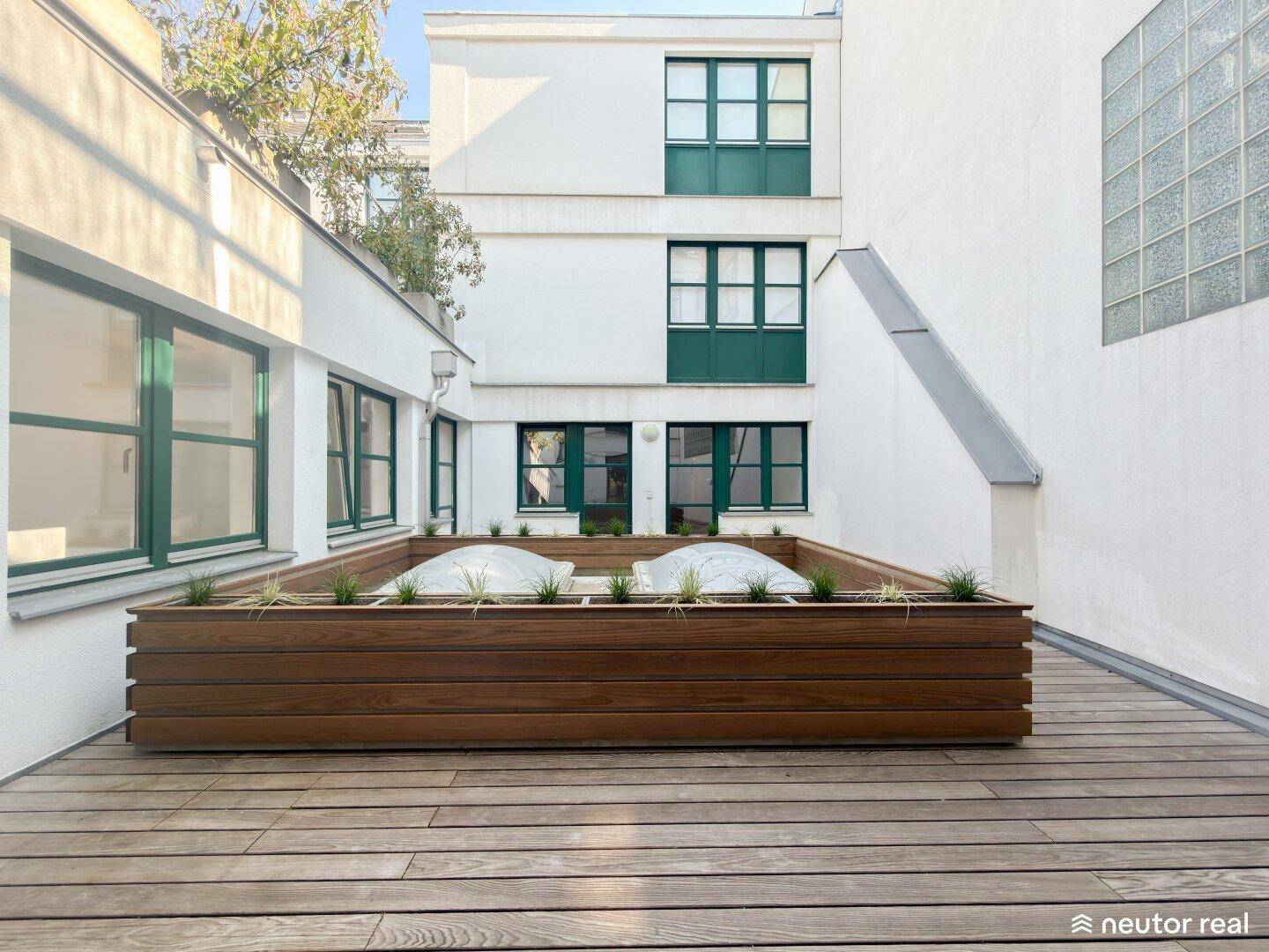 Sonniger Innenhof mit Holzterrasse und modernem Pflanzbeet, umgeben von Gebäuden.