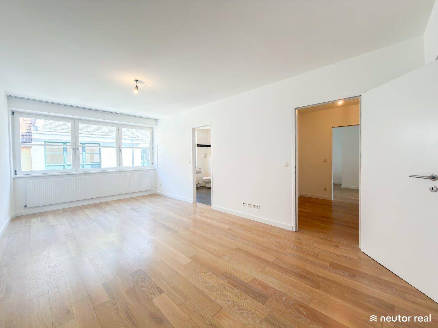 Helles Zimmer mit Parkettboden und Fenster, angrenzend an ein Badezimmer.