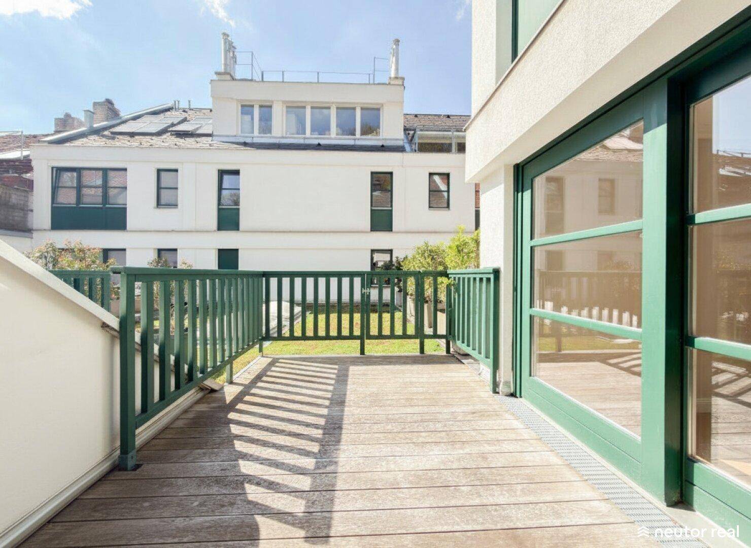 Geräumiger Balkon mit Holzbelag und grünen Geländern, bietet einen Blick auf die umliegenden Gebäude.