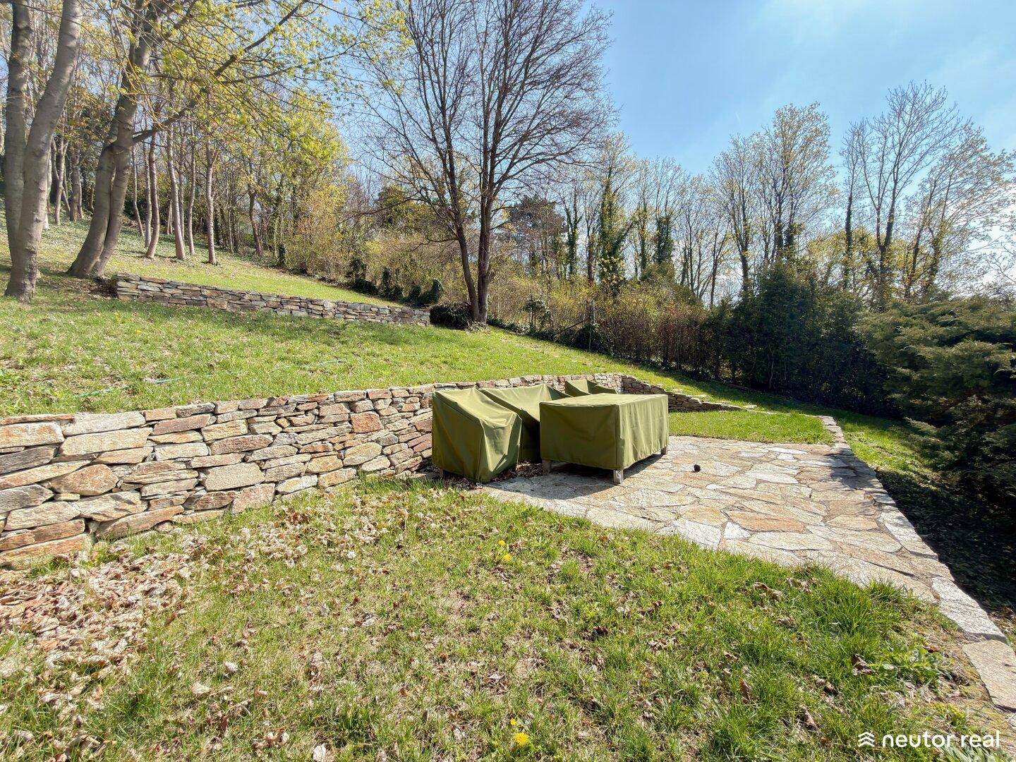 Gartenbereich mit Steinterrasse und abgedeckten Gartenmöbeln, umgeben von Rasen und Bäumen.