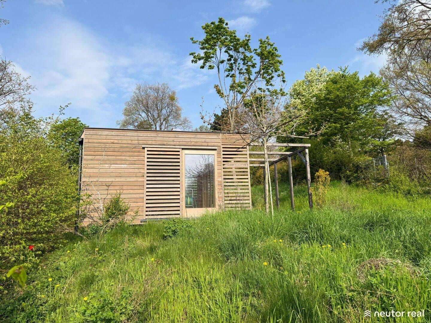 Modernes Holzhaus mit großen Fenstern und einer kleinen Pergola in natürlicher Umgebung.