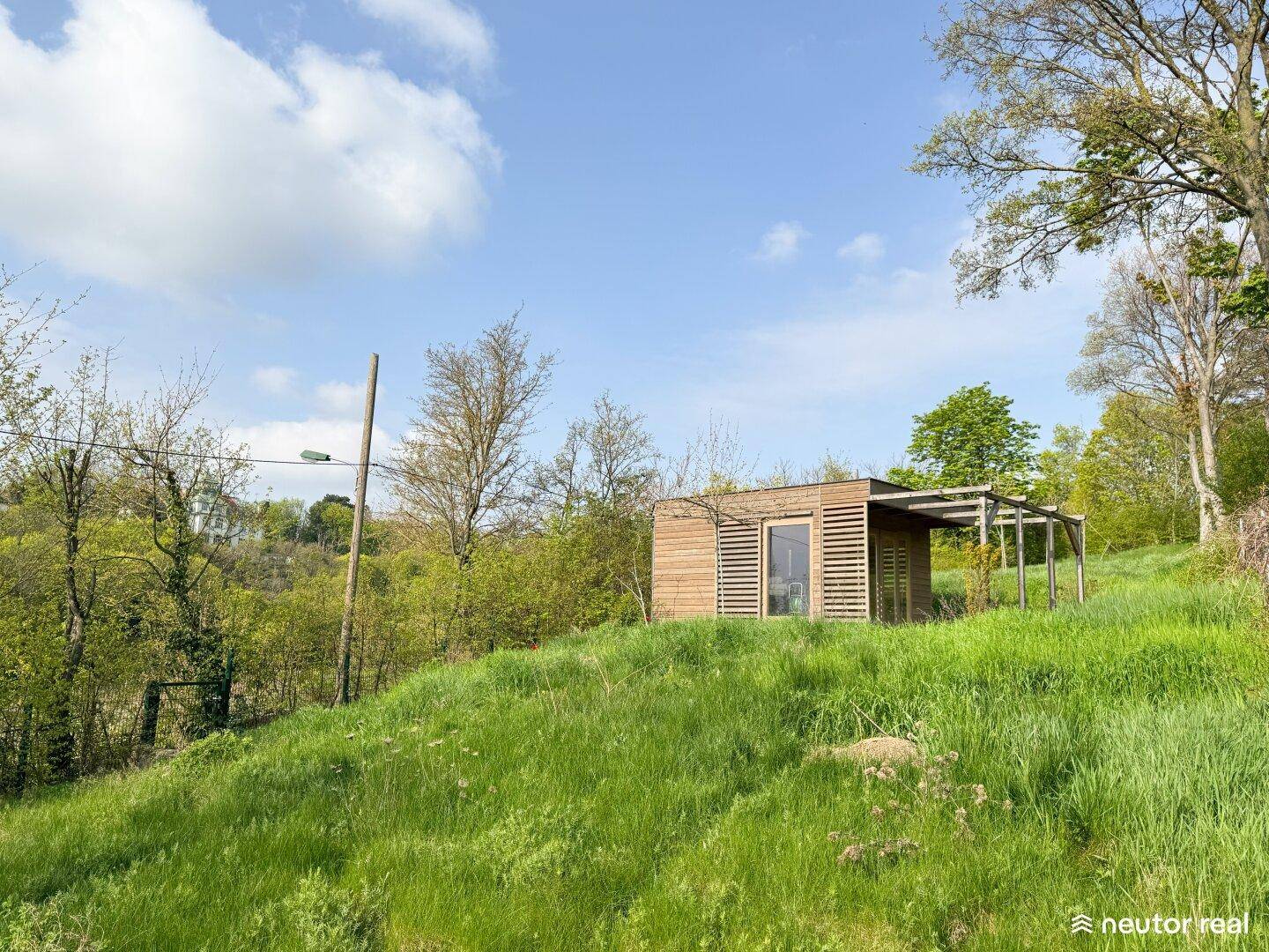 Kleines Holzhaus mit Lamellenfassade und Terrasse, umgeben von üppigem Grün und Bäumen.