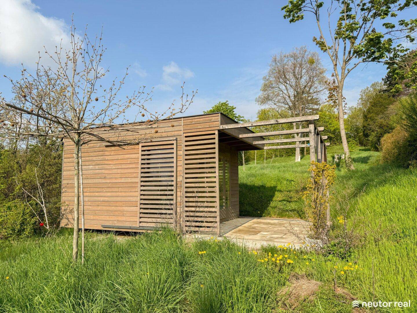 Kleines Holzhaus mit Pergola und großen Fenstern, umgeben von grüner Natur.