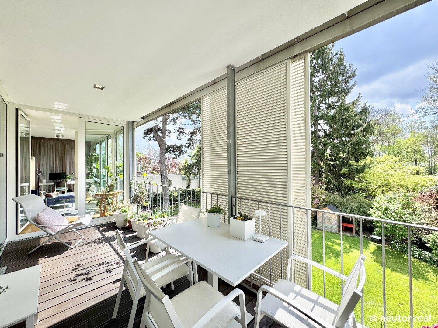 Heller Balkon mit Holzboden und Sitzgelegenheiten, umgeben von üppigem Grün und Bäumen.