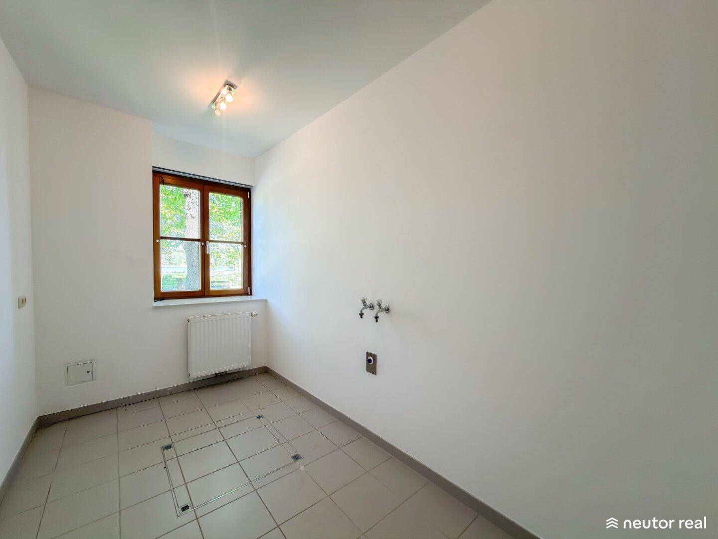 Leerer Raum mit Fliesenboden, Fenster und Anschlüssen für Waschmaschine oder Spüle, vielseitig nutzbar.
